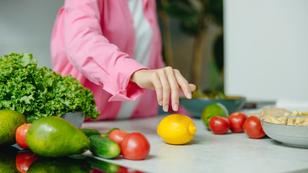 A person in a pink shirt reaching for a lemon surrounded by fresh vegetables.