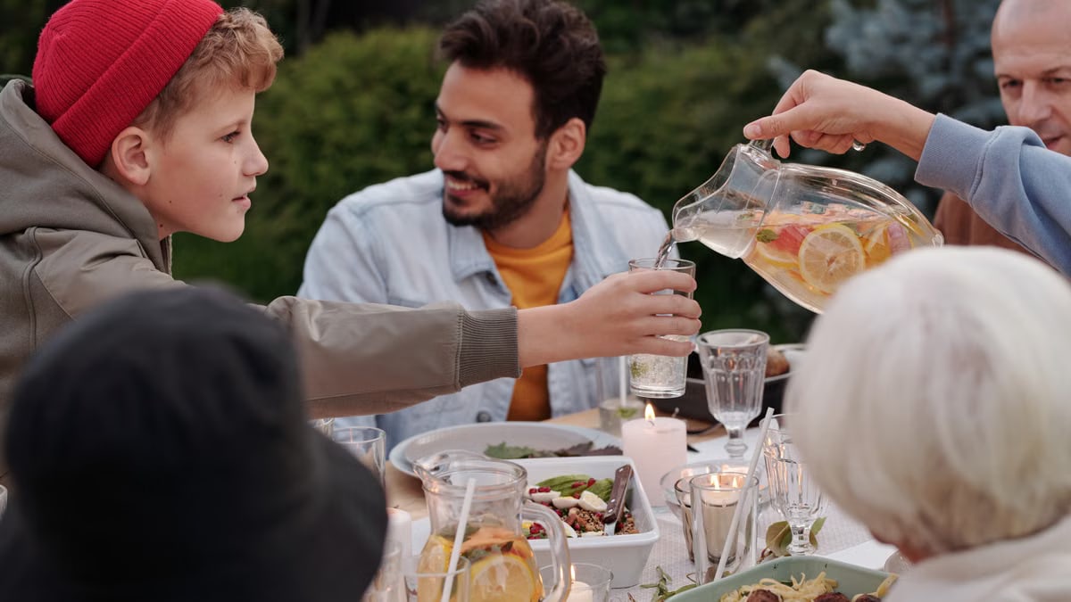 Faceless female pouring fruit beverage into glass of boy in warm cloth while cheerful multiethnic family having dinner outdoors